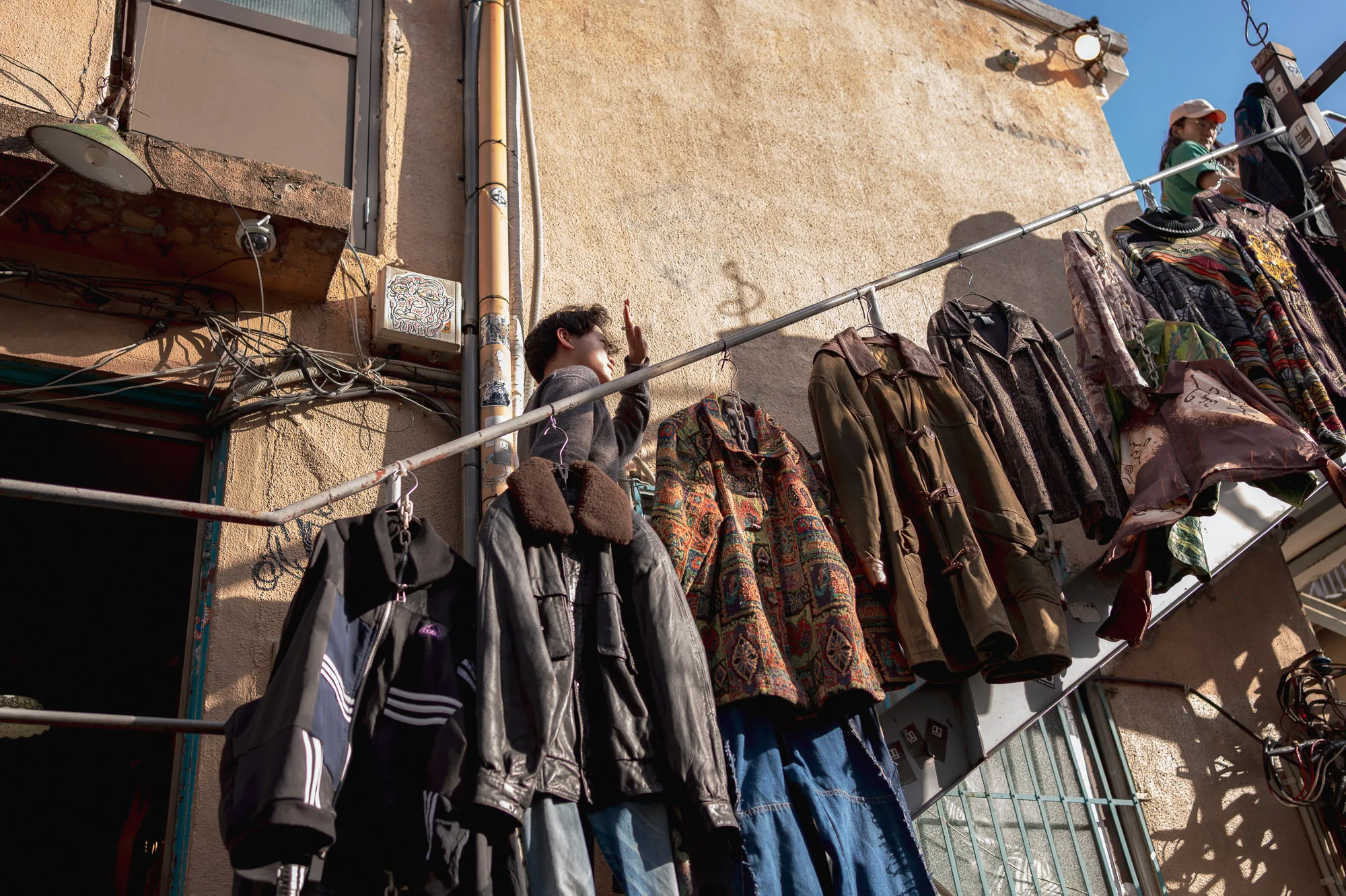 Street photography of vintage clothing displayed outside a shop in Shimokitazawa, Tokyo. Various jackets and clothing pieces hang on outdoor racks against a textured beige wall, capturing the authentic atmosphere of Tokyo's famous secondhand fashion district.