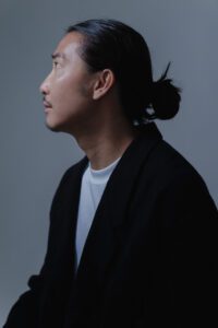 Editorial portrait of a male model with sleek black hair in a topknot, wearing a structured black blazer against a muted gray backdrop in JG Studio Tokyo. Dramatic side lighting accentuates angular features and hair texture, ideal for high-fashion portfolios or artistic campaigns.