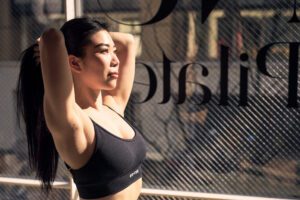 A female fitness model poses confidently, showcasing her toned physique in a stylish sports bra, with natural lighting highlighting her form against a minimalist backdrop at her pilates studio in Tokyo.