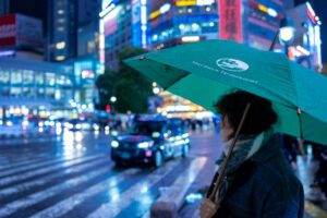 Lifestyle product photograph showcasing a green umbrella in the vibrant, neon-lit streets of Shibuya, Tokyo. This image captures the essence of urban nightlife, with reflections on the wet pavement and bustling city lights.