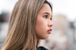 A model with long, golden-highlighted hair is captured in a soft profile shot, her gaze directed gently to the side. The image highlights her natural beauty and elegance, ideal for fashion and beauty portfolios. Shot on the balcony of JG Studio in Shibuya, Tokyo.