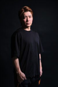 Portrait of Japanese DJ based in Tokyo, standing confidently in a black T-shirt against a dark backdrop. The image reflects the style and quality of JG Studio's photography services.