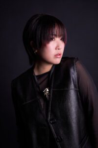 A stylish portrait of a woman with short hair and subtle pink highlights, wearing a sleek leather vest over a dark outfit, exuding confidence and elegance.