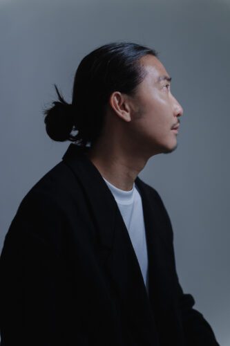Tokyo editorial portrait of a male model with sleek black hair in a topknot, wearing a structured black blazer against a muted gray backdrop. Dramatic side lighting accentuates angular features and hair texture, ideal for high-fashion portfolios or artistic campaigns.