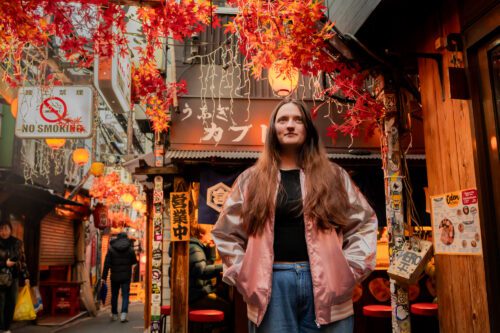 長い髪の若い女性が画像の中央に立ち、光沢のあるピンクのジャケットとブルージーンズを着ています。彼女は東京の新宿にある思い出横丁という狭い路地に位置しており、赤とオレンジの人工の葉や提灯などの秋の装飾で飾られています。背景には、日本語の文字が書かれた看板、「禁煙」の看板、そして独特の看板やメニューが表示された様々な居酒屋（日本のパブ）など、伝統的な日本の要素が特徴づけられています。路地は、歩いている人々や遠くに見えるいくつかの店で賑わっています。