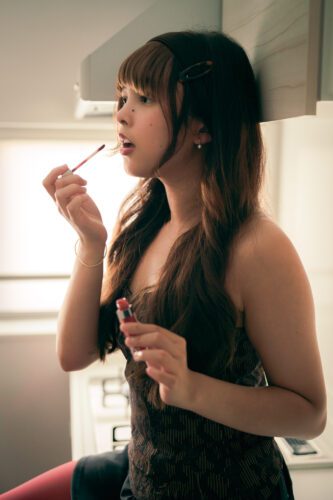 美容のクローズアップエディトリアル：劇的な映画的照明の中、東京のモデルが大胆な赤いリップスティックを塗る様子。メイクアップブランドやファッションキャンペーンに最適です。