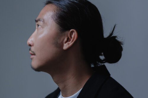 Profile Photo of a Chinese Artist Tokyo editorial portrait of a male model with sleek black hair in a topknot, wearing a structured black blazer against a muted gray backdrop. Dramatic side lighting accentuates angular features and hair texture, ideal for high-fashion portfolios or artistic campaigns.
