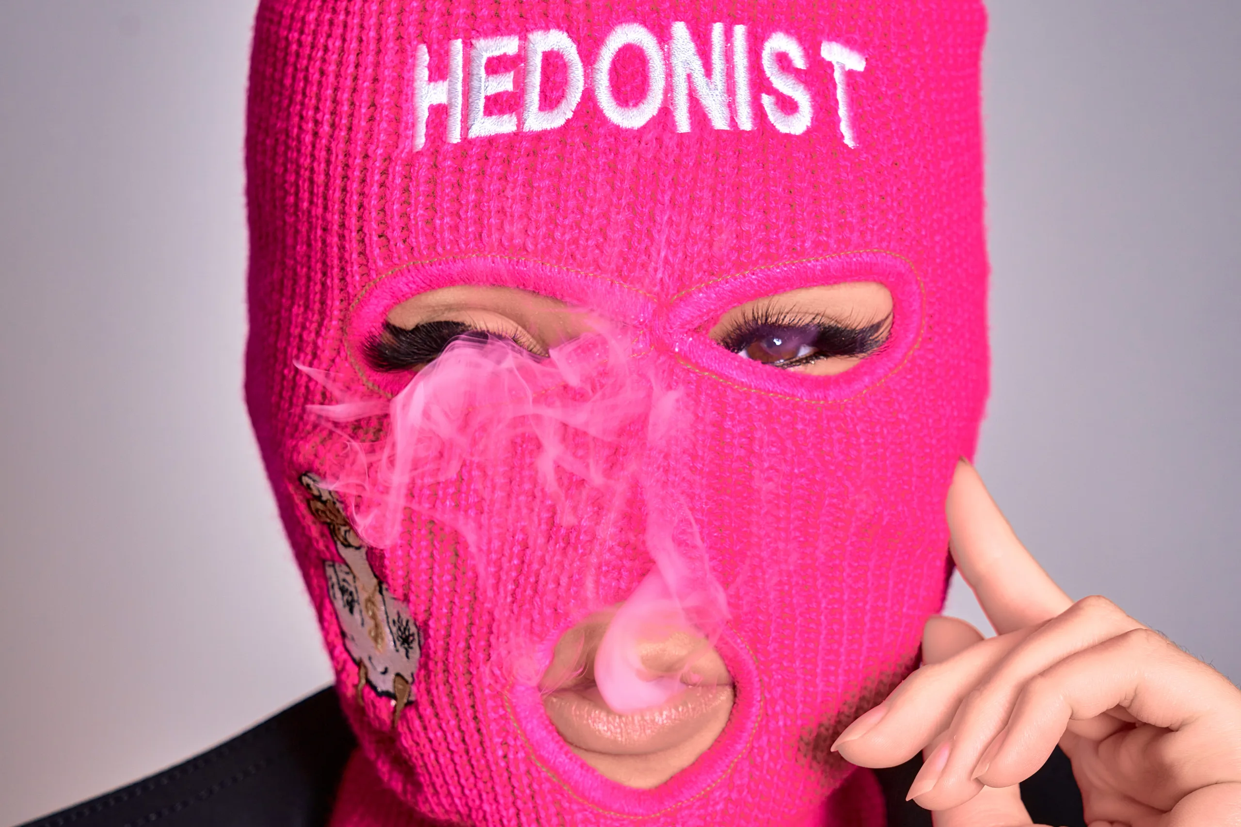 Japanese Brand Hedonist Woman wearing a pink balaclava with the word "Hedonist" and blowing smoke.