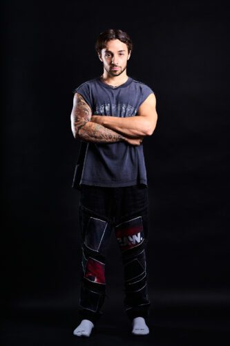 Fitness Photography A man with tattoed arms, standing with arms crossed, wearing a sleeveless shirt and patchwork pants against a dark background.