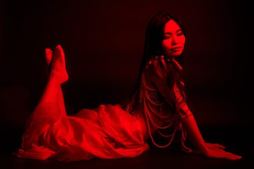 Colorful Portrait A woman in a flowing red dress adorned with beaded necklaces, sitting gracefully with her hands clasped, illuminated by a red light against a dark background.