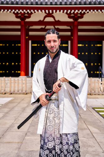 French Freestyle Footballer Séan Garnier A man dressed in traditional Japanese attire, holding a katana, standing confidently in front of a traditional Japanese temple with red pillars and golden accents.