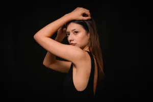 Portrait of a woman with long hair in a black sleeveless top, posing with arms raised, studio photography by JG Studio.