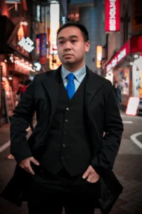 A businessman in a black suit and blue tie standing confidently in Tokyo’s vibrant nightlife district, captured in a professional corporate portrait by JG Studio.