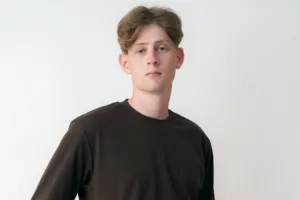 A young man poses for a studio portrait in a brown long-sleeve shirt against a white background. Professional headshot photography by JG Studio.