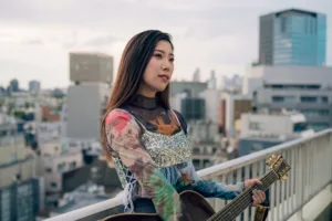 A young woman holds a guitar on a Tokyo rooftop, wearing a colorful floral outfit, with the cityscape in the background. Lifestyle portrait photography by JG Studio.