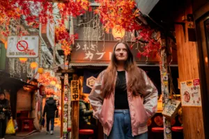 Person posing in a Tokyo alley decorated with autumn leaves and lanterns, wearing a shiny pink jacket.
