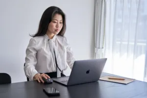 Focused Professional Work Business woman sitting at a dark wood desk using a laptop and mouse in a bright, minimalist office.