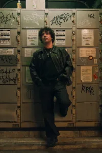 A man in a black leather jacket and dark jeans leaning against a wall of metallic coin lockers covered in graffiti in a Shibuya alleyway at night.