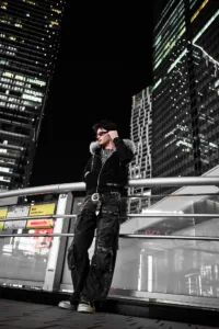 A young man posing for a professional portrait at night in Shibuya, Tokyo. He is wearing a black fur-lined hoodie, dark cargo pants, and sunglasses, leaning against a silver railing with illuminated skyscrapers in the background.