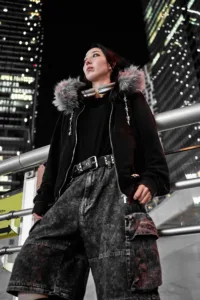 A woman in a black fur-lined hoodie and oversized denim shorts posing at night against a backdrop of illuminated Shibuya skyscrapers.