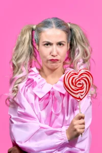 A high-fashion portrait of a woman with silver-blonde pigtails and a moody expression, wearing a ruffled pink blouse and holding a large red-and-white heart-shaped lollipop against a vibrant pink background.