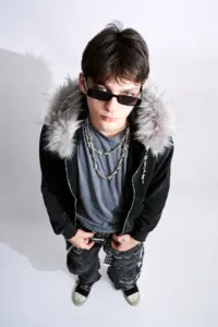 High-angle studio portrait of a model wearing a Post Xeno faux-fur hooded jacket, barbed wire necklaces, and distressed streetwear by Tokyo photographer JG Brasseur.