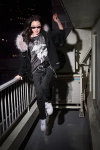 Post Xeno Streetwear Campaign - Fashion Photography Model wearing a Post Xeno streetwear outfit on a Shibuya balcony at night, featuring a fur-lined hoodie, graphic t-shirt, and studded belt.