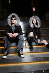 Two models sitting on illuminated stairs in Tokyo wearing Post Xeno streetwear, featuring black oversized hoodies with large faux-fur trimmed hoods, distressed denim cargo pants, and high-top sneakers.