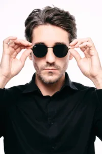 A high-contrast studio portrait of JG wearing round black Rayban sunglasses and a dark button-down shirt against a seamless white background. The lighting is bright and clean, emphasizing a professional and modern aesthetic for fashion photography in Tokyo.