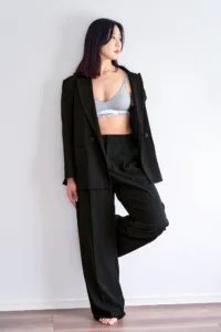 A professional fashion portrait of a woman in a black power suit and grey Calvin Klein bralette, posing against a white wall at JG Studio in Tokyo.