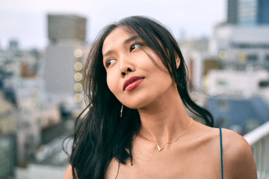 A professional portrait of a woman with dark hair and red lipstick, wearing a gold V-shaped necklace, captured against a soft-focus urban background in Tokyo at dusk.