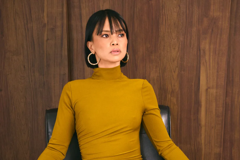 Close-up of a model in a yellow dress with gold hoop earrings against a warm wooden backdrop.