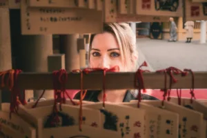 東京の神社で、木製の絵馬の隙間からこちらを見つめる女性を捉えた、クリエイティブなクローズアップポートレート。