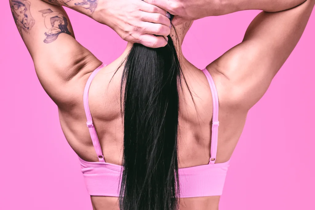 Rear close-up of fitness model Meru tying her long black hair, showcasing toned shoulders and tattoos against a bright pink studio background, by JG.