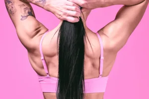 Rear close-up of fitness model Meru tying her long black hair, showcasing toned shoulders and tattoos against a bright pink studio background, by JG.