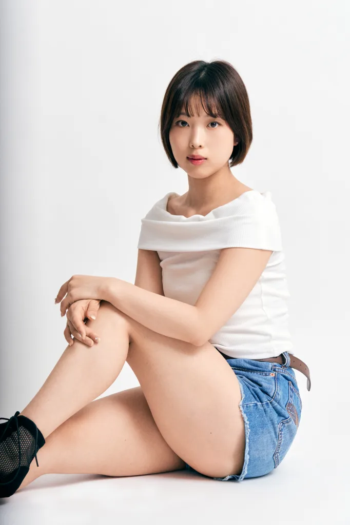 東京でのスタジオポートレート撮影。白背景を背に、ショートボブの女性が座り、白いオフショルダートップとデニムショートパンツを着用。柔らかいスタジオ照明。