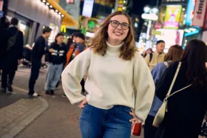 Smiling woman in a white turtleneck posing on a busy Shibuya street at night, surrounded by neon signs, during JG's Shibuya photo tour.