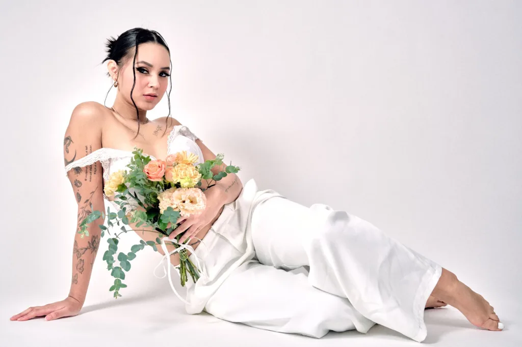 Model in white reclining on the studio's white backdrop with a bouquet of flowers, natural light.