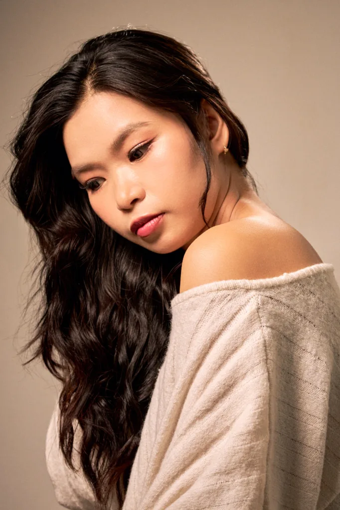 Beauty portrait in a Tokyo studio: long dark waves, an off-the-shoulder cream knit and a lowered gaze against a warm beige backdrop.