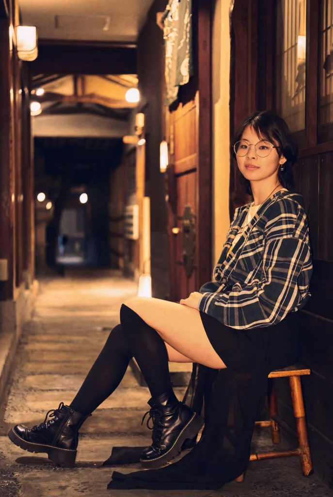 Evening portrait in a lantern-lit wooden alley in Tokyo: a woman in a plaid shirt, black skirt and boots, seated on a low wooden stool.