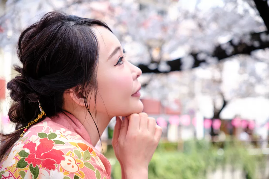 Profile portrait of a woman in a pink floral kimono looking up under cherry blossoms, chin resting on her hands, pink lanterns blurred behind her.