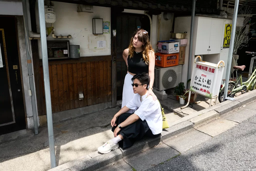 Outdoor portrait session in Shimokitazawa by day: a couple, she standing with her hand on his shoulder, he seated on the kerb, in front of an old shopfront, Tokyo, by JG.