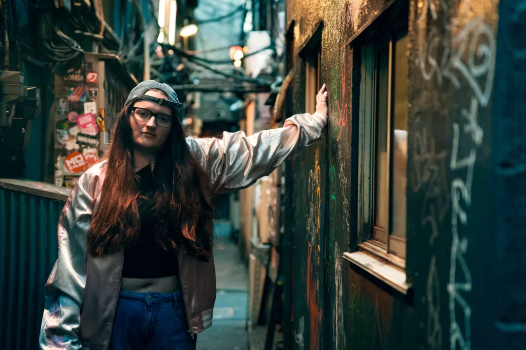Outdoor portrait session in Shinjuku by night: a woman with long auburn hair in a backward cap and glasses, wearing a satin bomber jacket, her hand on a graffiti wall in a narrow alley covered in stickers and cables, Tokyo, by JG.