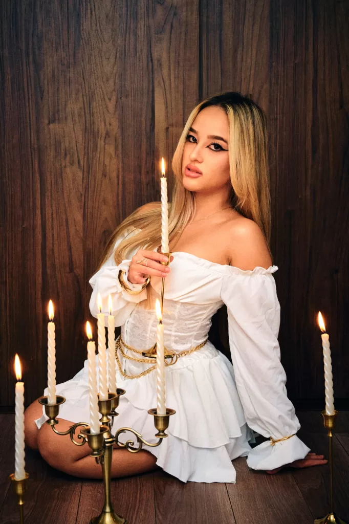 Candlelit studio portrait of a woman with long blonde hair in a white off-the-shoulder dress, holding a lit candle among brass candlesticks, dark wood backdrop, by JG.