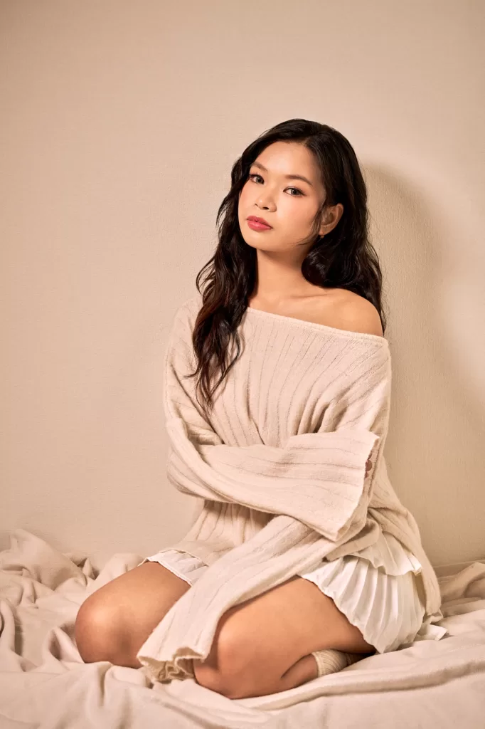 Studio portrait of a woman with long dark waves in an off-the-shoulder cream knit, kneeling on a blanket against a warm beige backdrop, Tokyo, by JG.