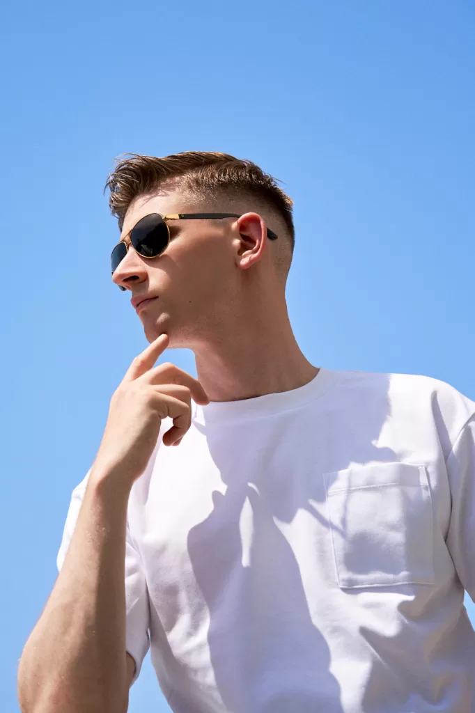 Low-angle portrait of a man in gold-framed sunglasses and a white pocket t-shirt, hand at his chin, on the studio balcony in Shibuya against a clear blue sky, by JG.