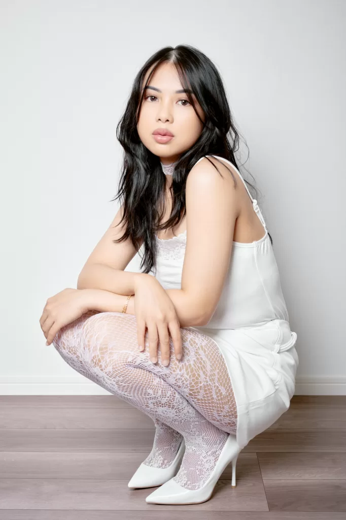Studio portrait of a woman crouching in a white camisole, white lace tights and white heels against a white wall at the Shibuya studio, by JG.