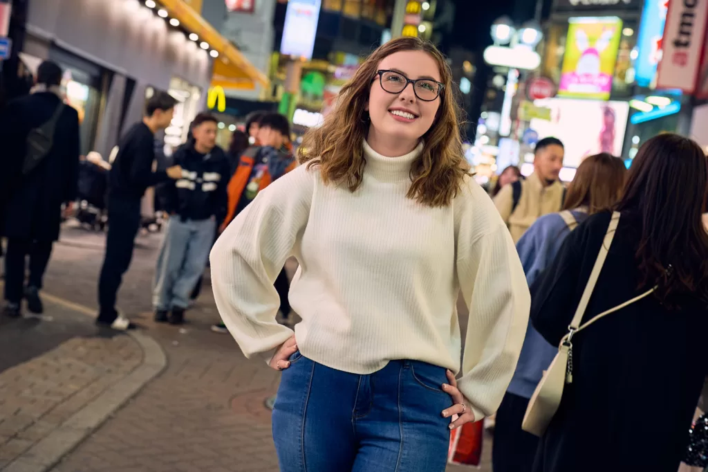 夜の渋谷、ネオンが輝く賑やかな通りでのポートレート。黒縁の眼鏡にクリーム色のリブ編みタートルネック姿の女性が、両手を腰に当てて微笑み、上を見上げる。背景に行き交う人々と光る看板。東京にてJG撮影。