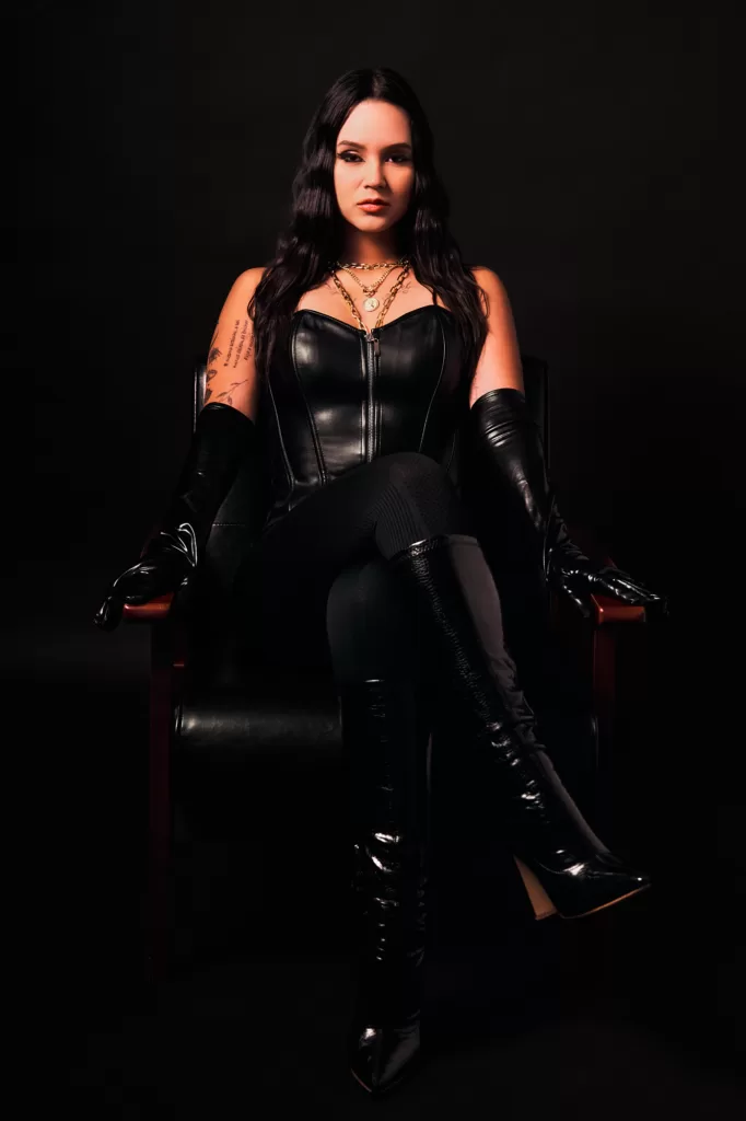 黒髪のロングウェーブの女性が、黒いレザーのコルセットに重ねづけしたゴールドのチェーン、黒のロンググローブ、膝丈のエナメルブーツ姿で、脚を組んで革のアームチェアに座るスタジオポートレート。黒い背景。東京にてJG撮影。