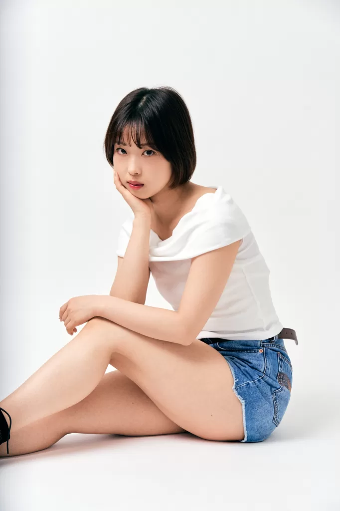 ショートボブの女性が、白いオフショルダーのトップスとデニムのショートパンツ姿で床に座り、手にあごをのせるスタジオポートレート。白い背景。東京にてJG撮影。